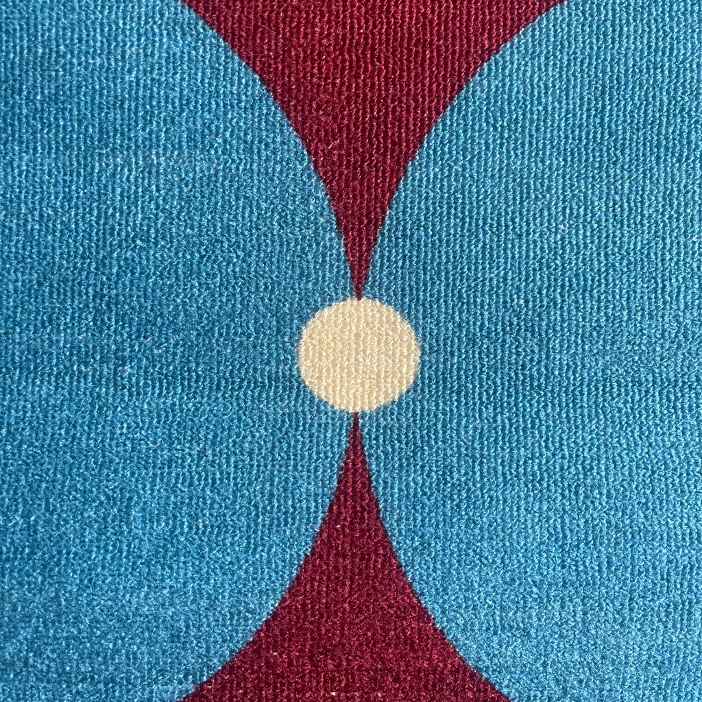 A close-up of a textured fabric or rug featuring a mid-century modern geometric pattern with large blue and burgundy shapes meeting at a central cream circle.